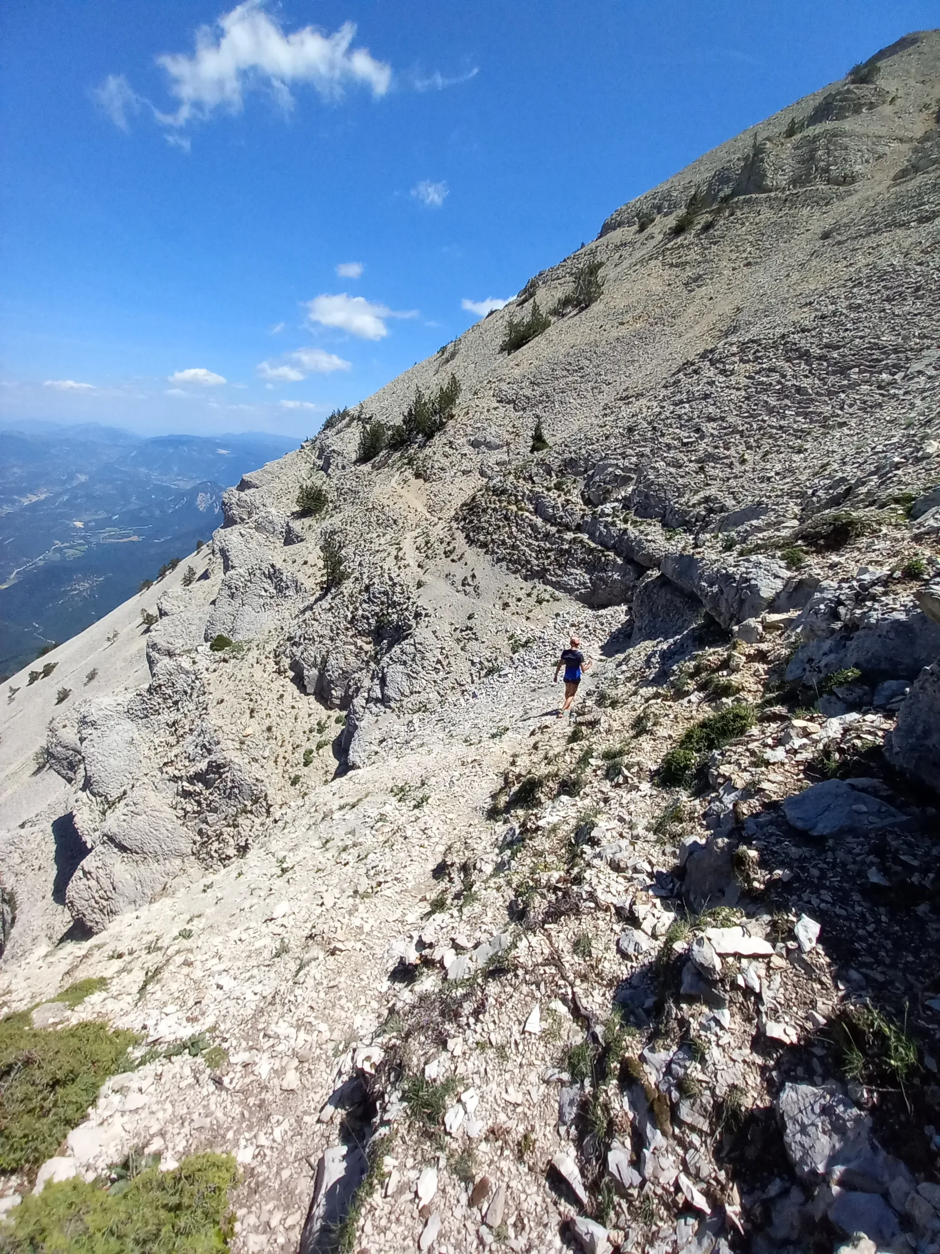Un coureur seul progresse sur un pierrier, à flanc de crête calcaire, très au-dessus d'une vallée