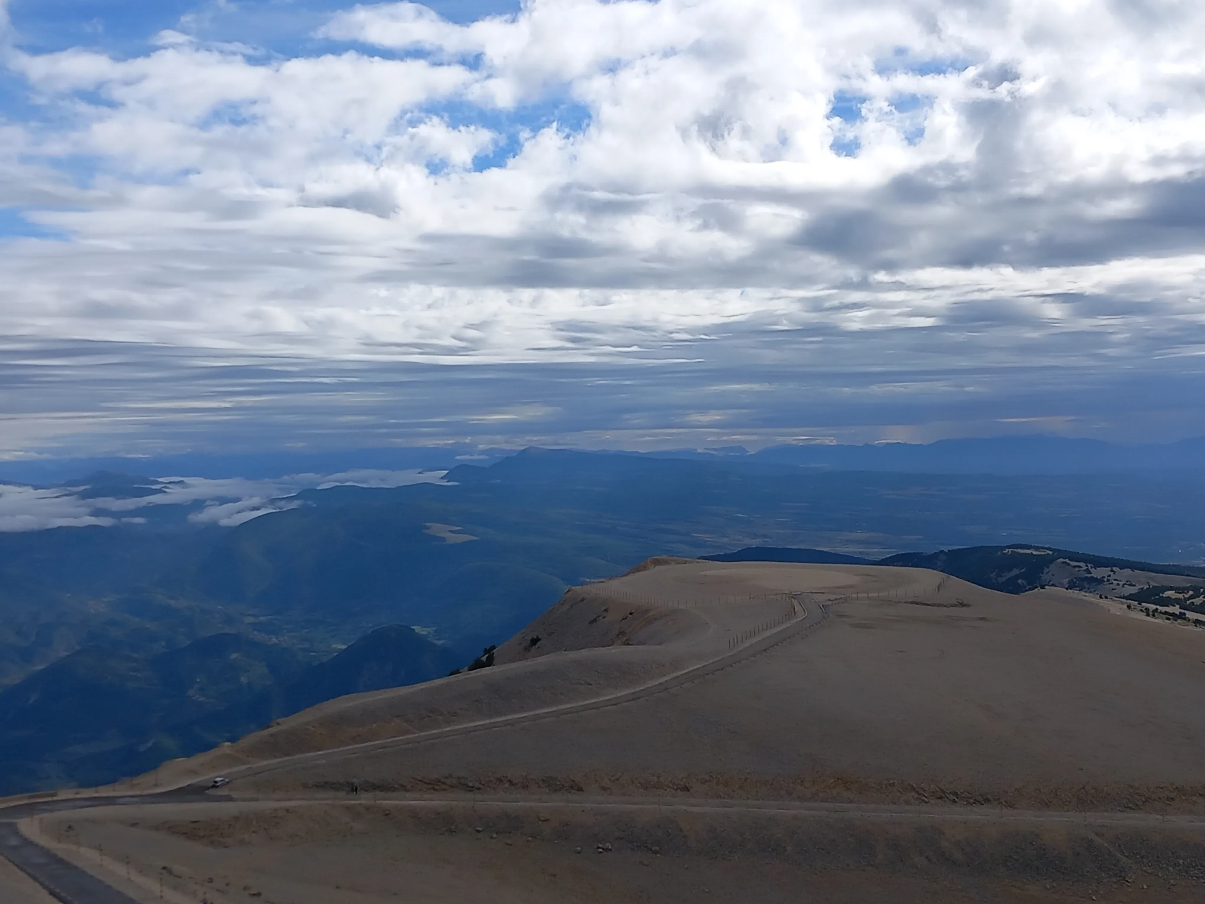 Le sommet dénudé et caillouteux d'une montagne, une route qui en suit la crête, et une mer de nuages sur les reliefs au loin