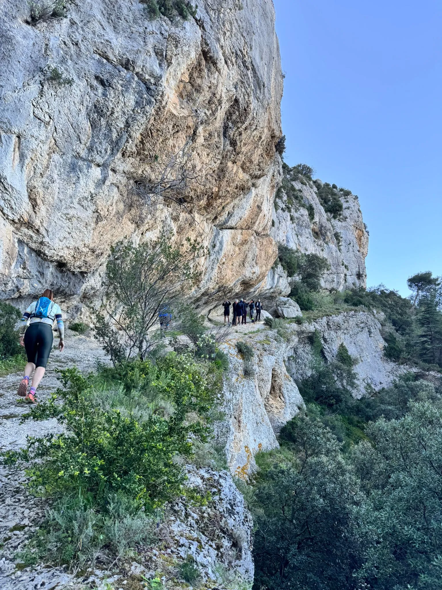 Une coureuse remonte un sentier de pierre au pied d'une haute paroi calcaire, et un groupe s'est arrêté plus loin sous le surplomb