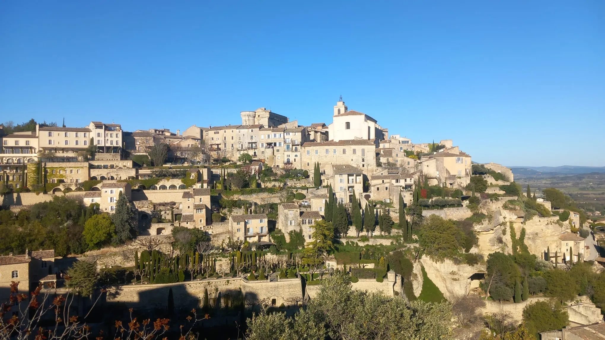 Un village de pierre perché en terrasses au-dessus d'une falaise, avec son clocher, ses cyprès et la plaine au loin